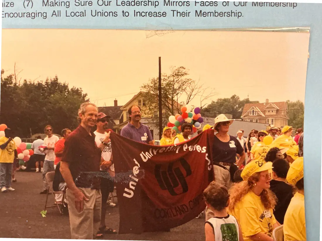 Color photograph of marchers on a street holding a dark red union banner with partly visible lettering; people in yellow caps and balloons in the background.