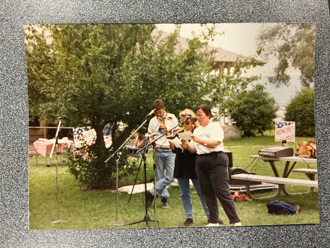 Color photograph: two musicians with guitar and fiddle at microphones on a lawn; a red, white and blue Hinchey campaign sign on a table.
