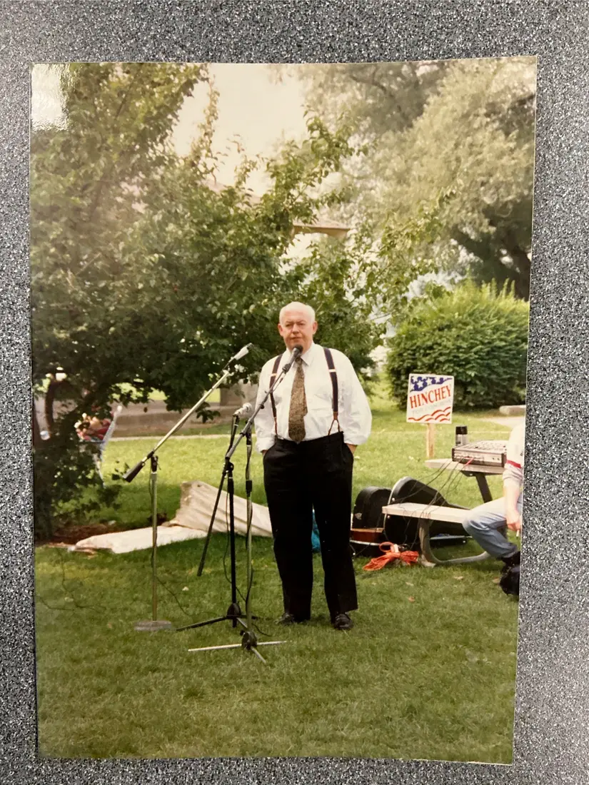 Color photograph: an older man in a white shirt, tie, and suspenders standing at a microphone on a lawn, with a Hinchey sign behind him.