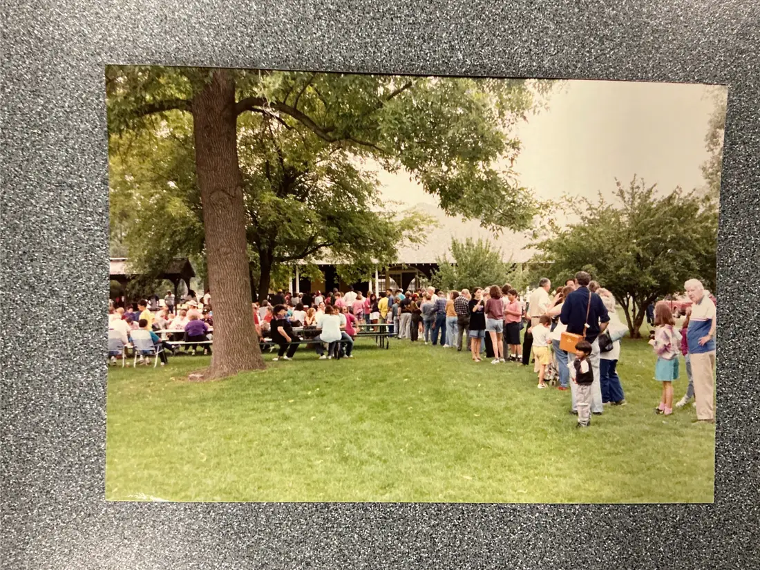 Color photograph: a crowd of people standing and sitting under large trees at an outdoor gathering.