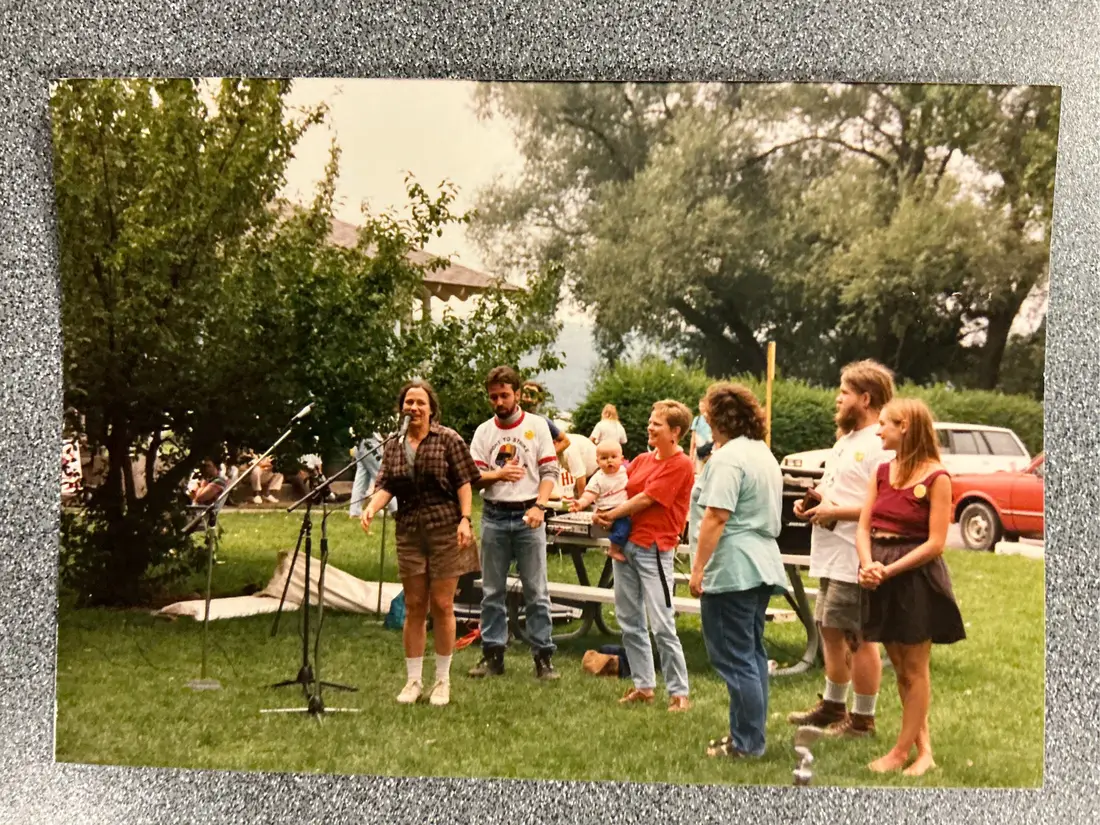 Color photograph: people standing around picnic tables on grass beside a parked red car.