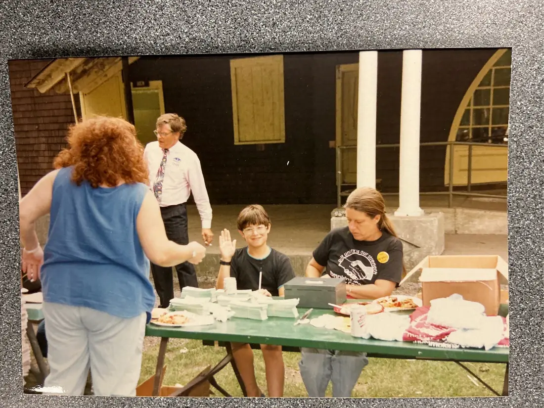 Color photograph: people serving and collecting food at a long table outside a building.