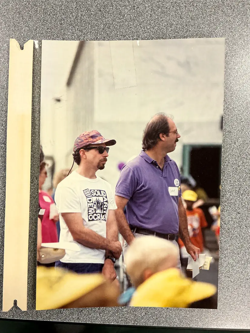 Color photograph of two men at a crowded outdoor event; one in a cap and white T-shirt printed with the words Solidarity and Democracy, the other in a purple polo shirt with a name tag.