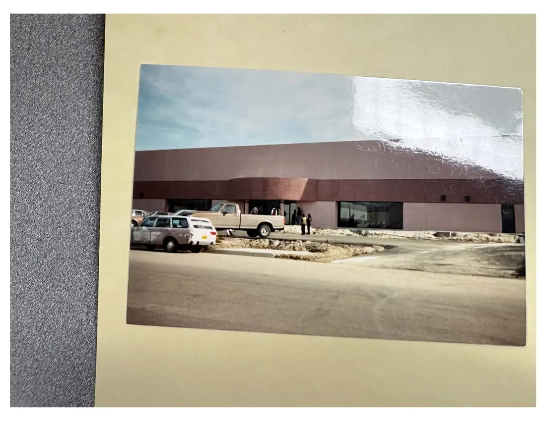 Color snapshot of a long, low industrial building with a brown roof and a parking lot with cars in front.