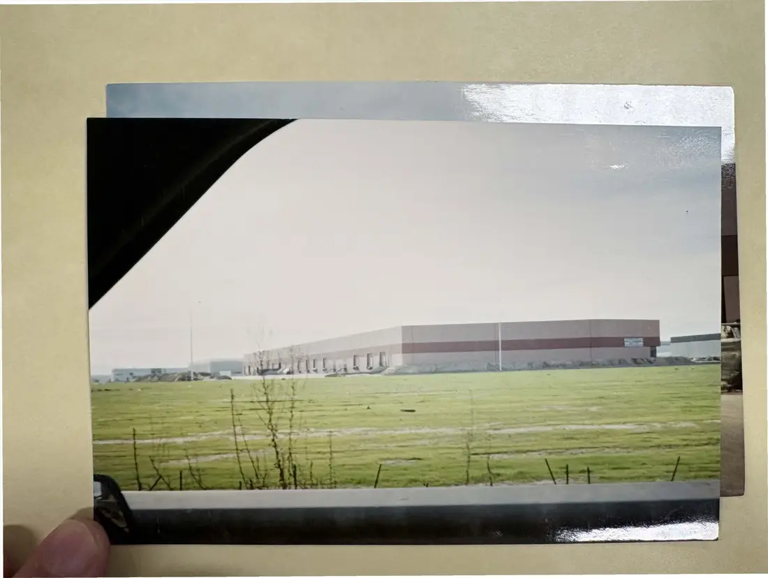 Color snapshot, held in a hand, of a large industrial building beyond a green field.